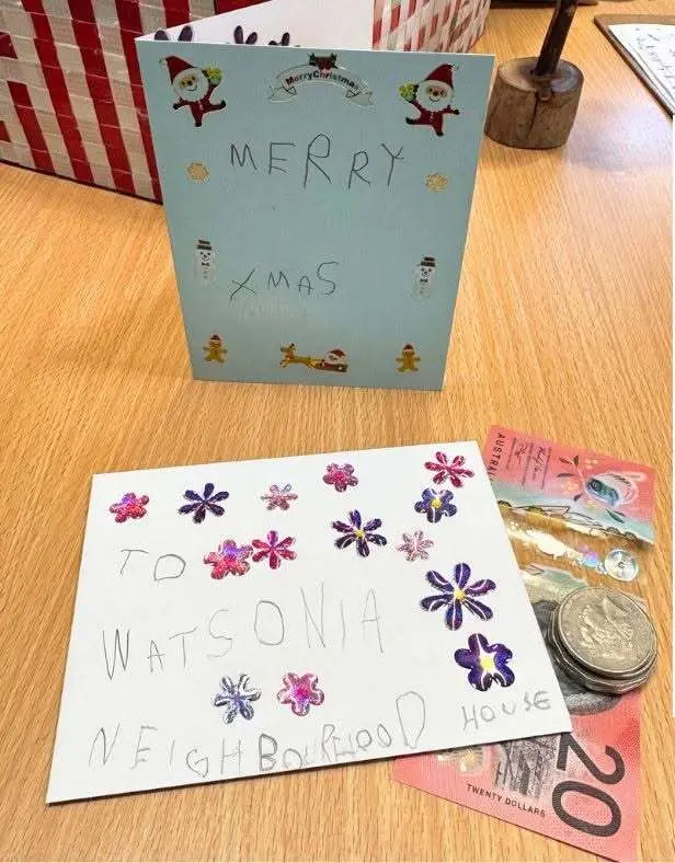 Two handmade cards on a table — one saying Merry Xmas with drawn decorations, one saying To Watsonia Neighbourhood House decorated with paper flowers — beside coins and a twenty dollar note