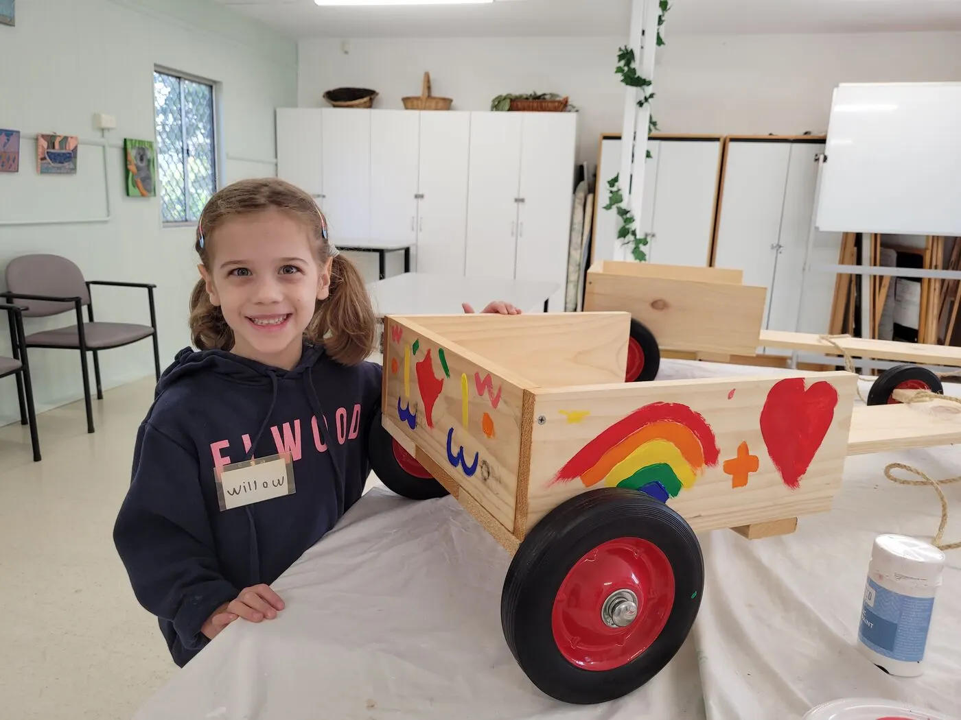 A child standing proudly beside a finished billy cart painted with a rainbow and a heart