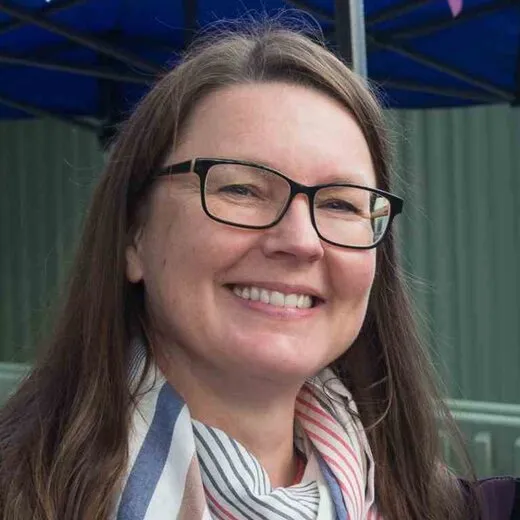 Lucie Holden smiling, wearing glasses and a scarf, outdoors at the House