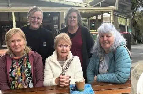 Five people gathered around a table outside a cafe with coffees, smiling at the camera