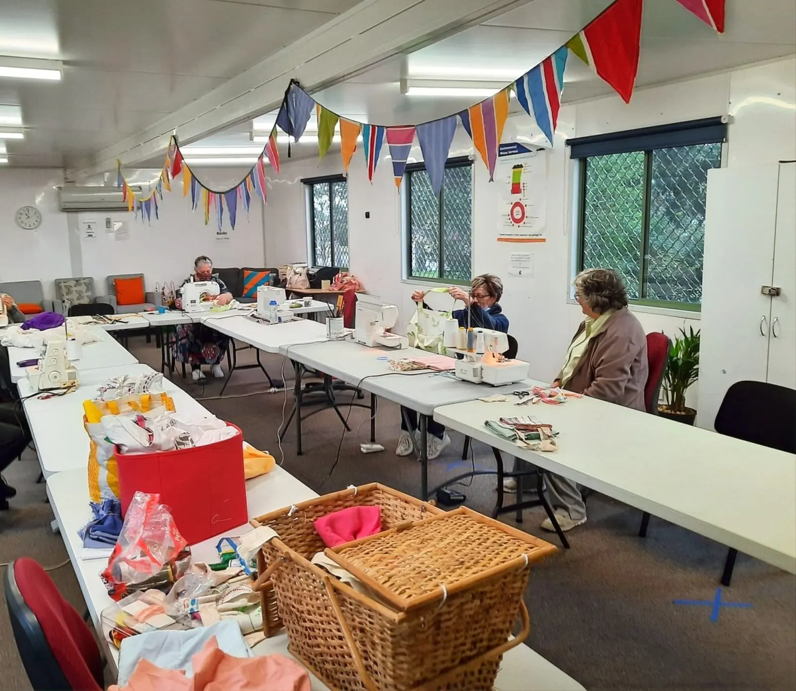 Long tables covered with craft materials and baskets, a few people working, bunting strung overhead